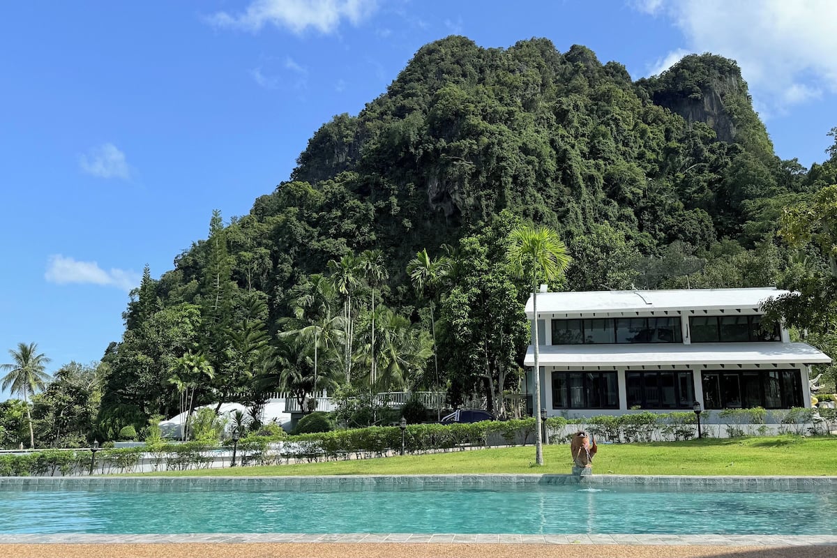 Um edifício moderno com piscina em frente, cercado por vegetação exuberante e montanhas ao fundo.
