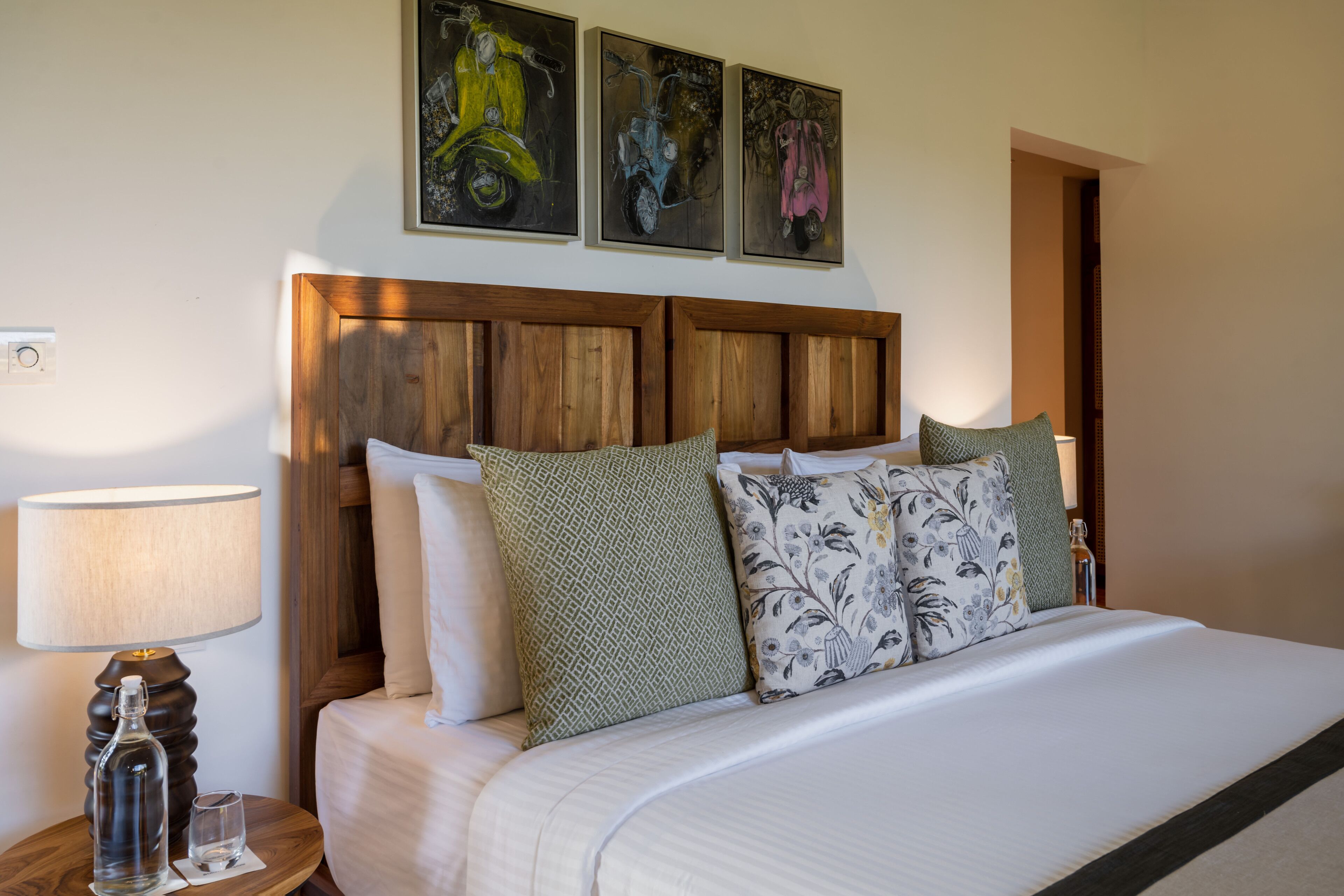 A neatly made bed with patterned pillows, a wooden headboard, and a wall-mounted artwork of scooters.