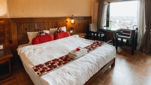 Habitación de hotel con una cama grande, vigas de madera, ventilador de techo, ventana con cortinas y vistas a la ciudad.