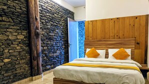 A bedroom with a stone wall, wooden headboard, and a bed with white bedding and two orange pillows. - Gantavya Resort (Pokhara)