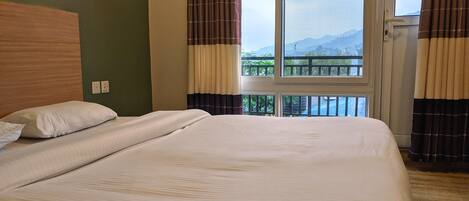 A hotel room with a large bed, a wooden headboard, a window with curtains, and a view of mountains.