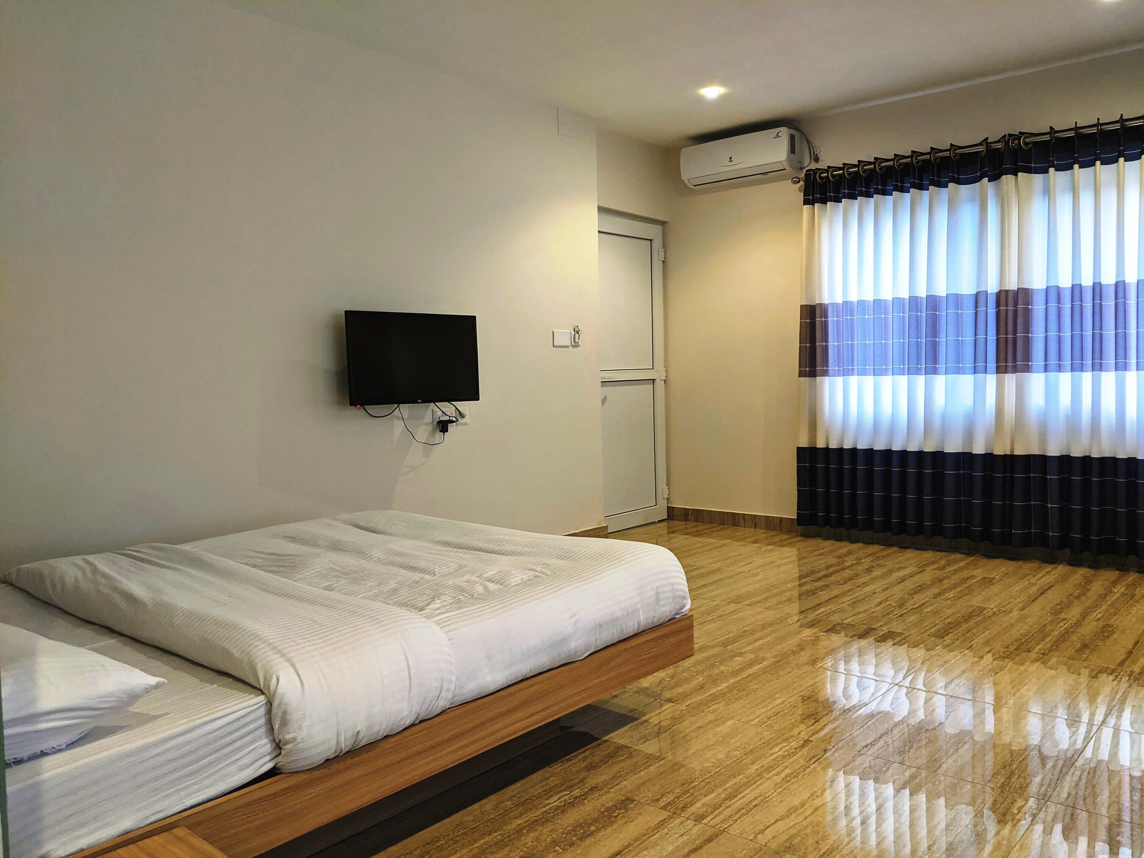 A hotel room with a bed, a flat-screen TV mounted on the wall, an air conditioning unit, and a window with striped curtains.