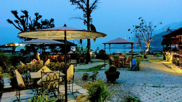 An outdoor seating area with tables and umbrellas, surrounded by trees and a view of the sea. - Gantavya Resort (Pokhara)