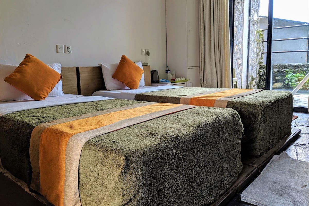 Een hotelkamer met twee bedden, een raam met gordijnen en uitzicht op groen, en een klein tafeltje met een lamp.