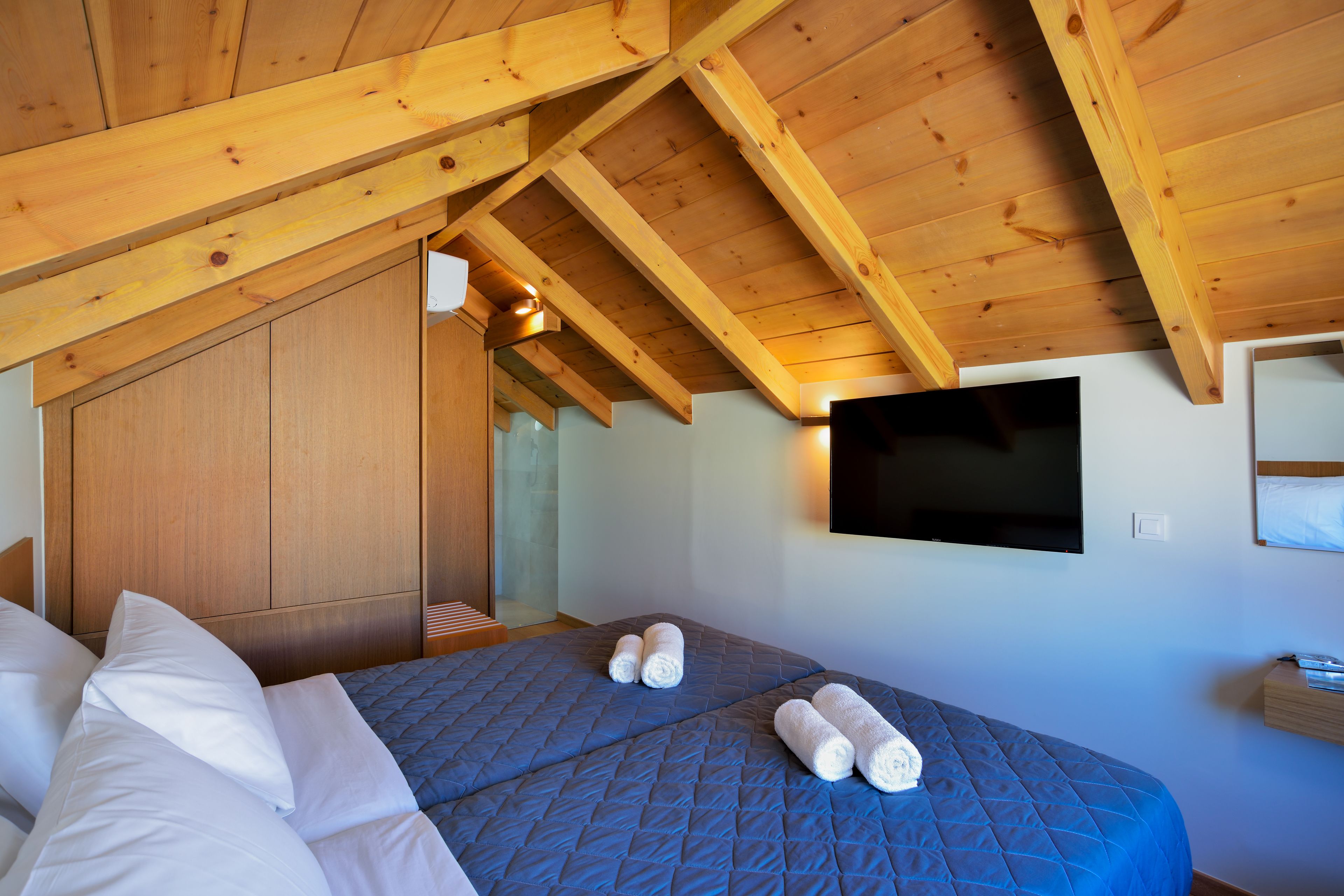 A bedroom with a sloped ceiling, wooden beams, a bed with blue bedding, white pillows, and two rolled towels.
