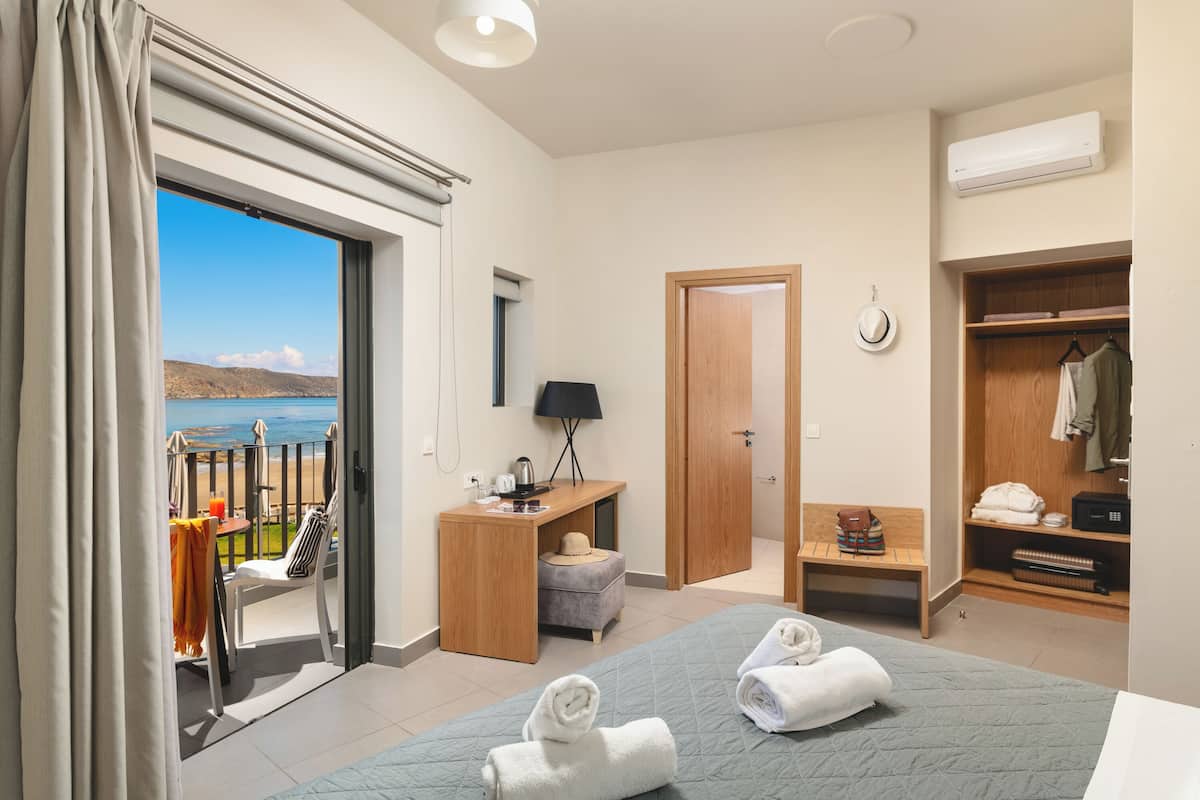 Habitación de hotel con cama, escritorio, silla, mesita, banco, armario y vista a la playa.
