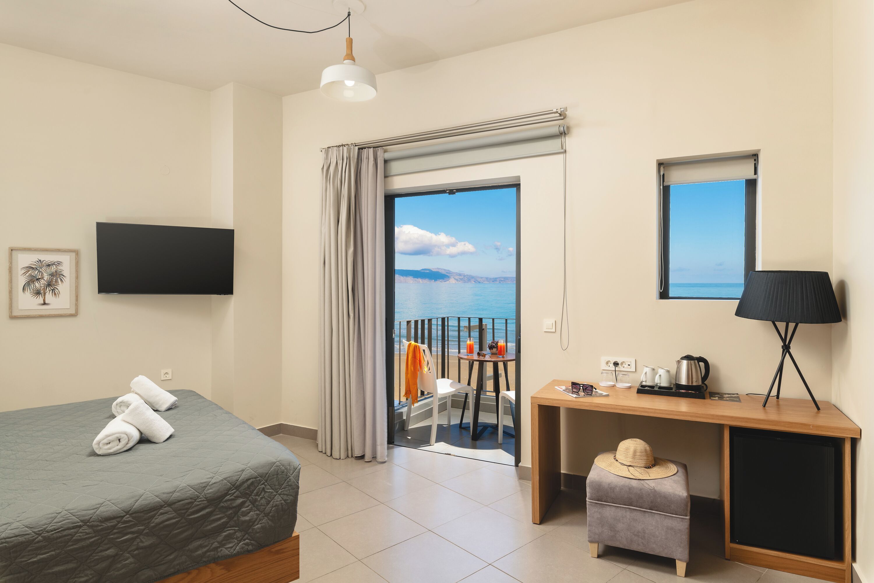 A hotel room with a bed, a desk, a chair, a small table with a lamp, a television, and a view of the sea.