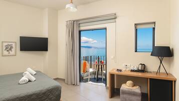 A hotel room with a bed, a desk, a chair, a small table with a lamp, a television, and a view of the sea.