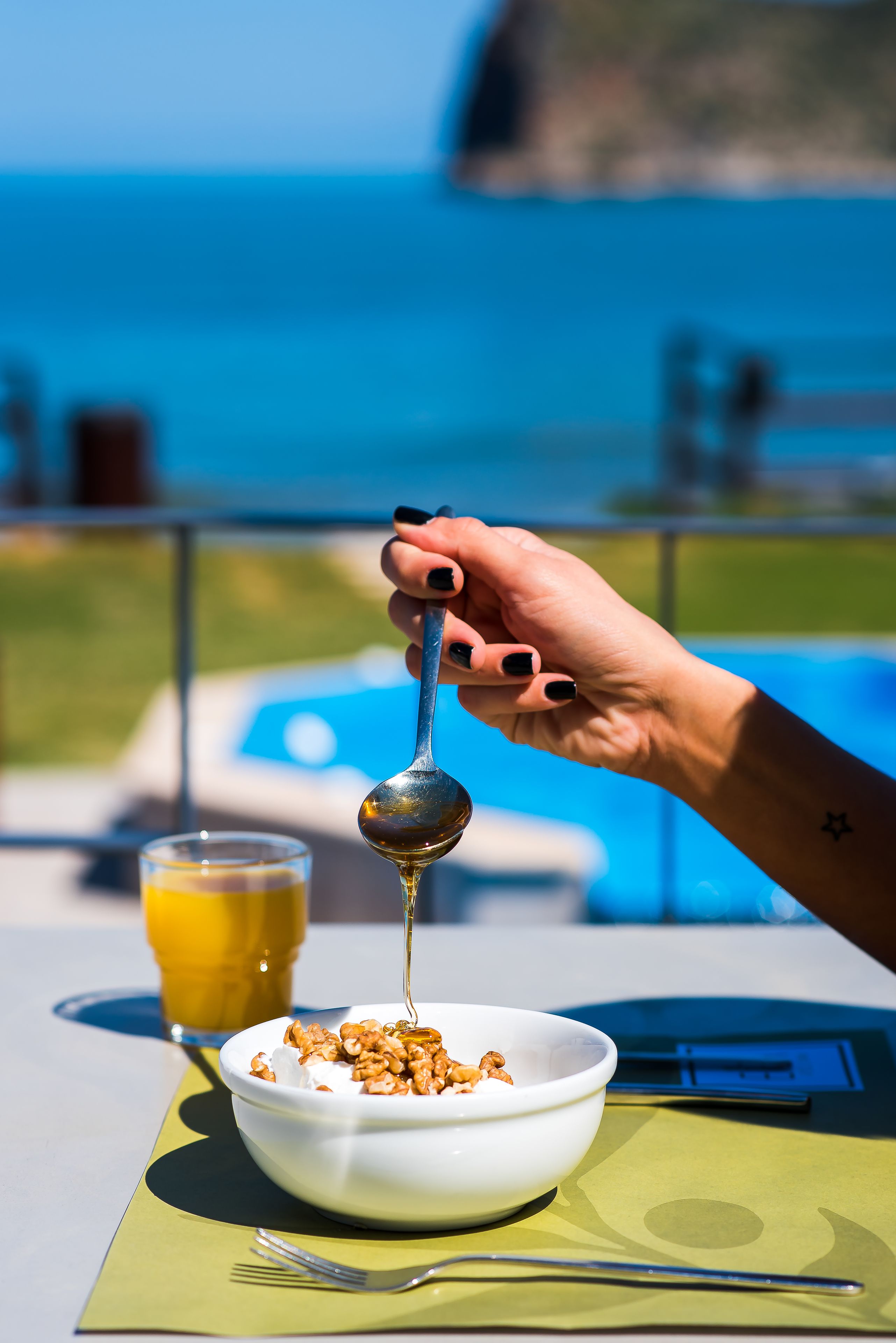 A hand holding a spoon over a bowl of cereal with milk, a glass of orange juice, and a poolside view in the background.