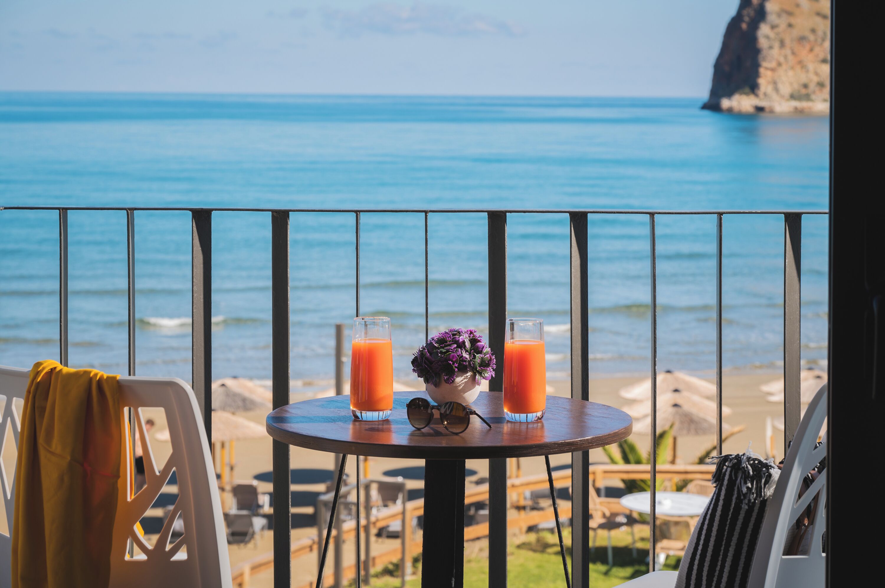 Un balcon avec une table sur laquelle se trouvent deux verres de jus d’orange, une paire de lunettes de soleil et une petite fleur violette. La vue comprend une plage et la mer.