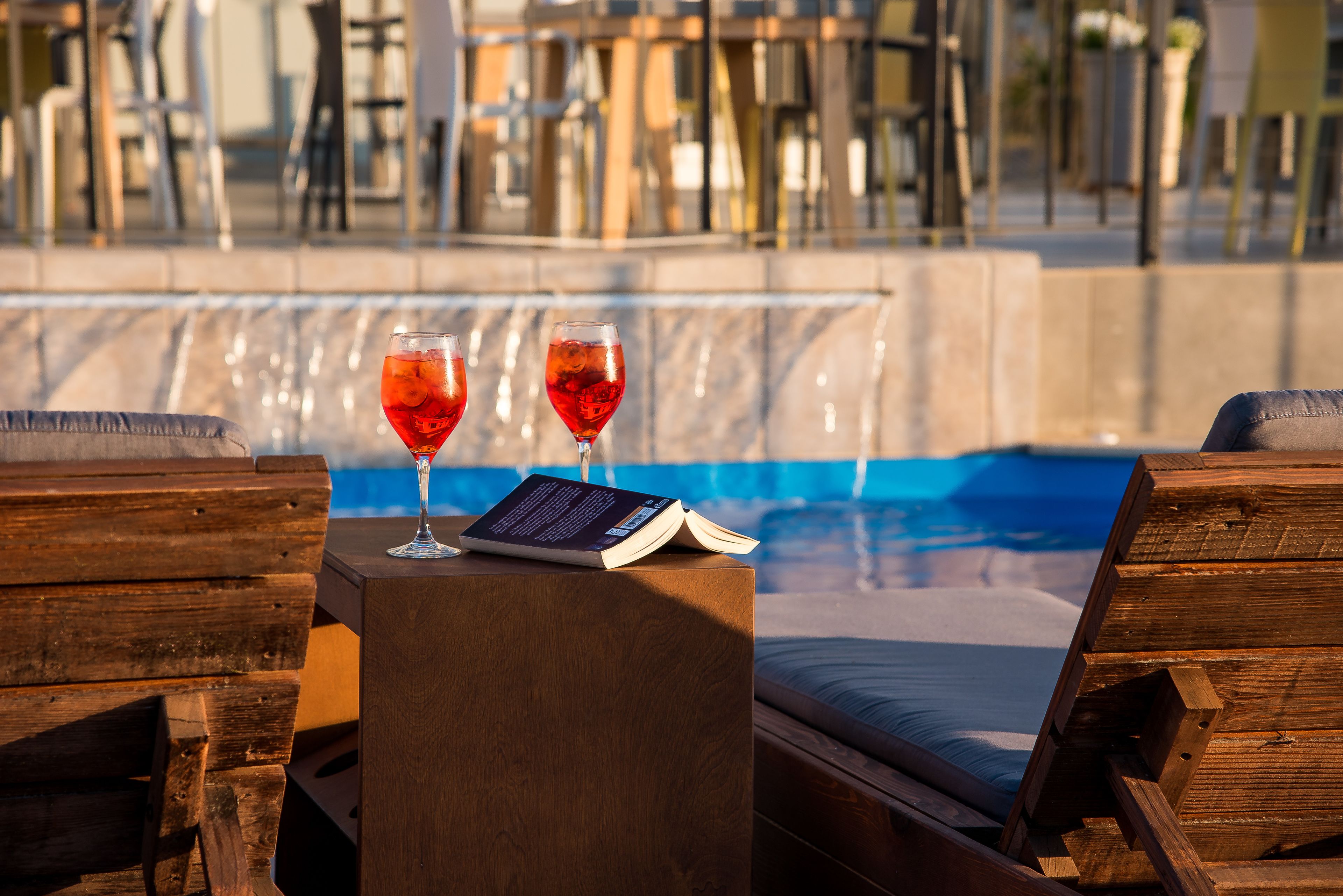 A poolside area with wooden loungers, a small table with two glasses of red drink and a book.