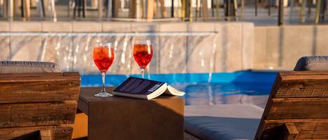 A poolside area with wooden loungers, a small table with two glasses of red drink and a book.