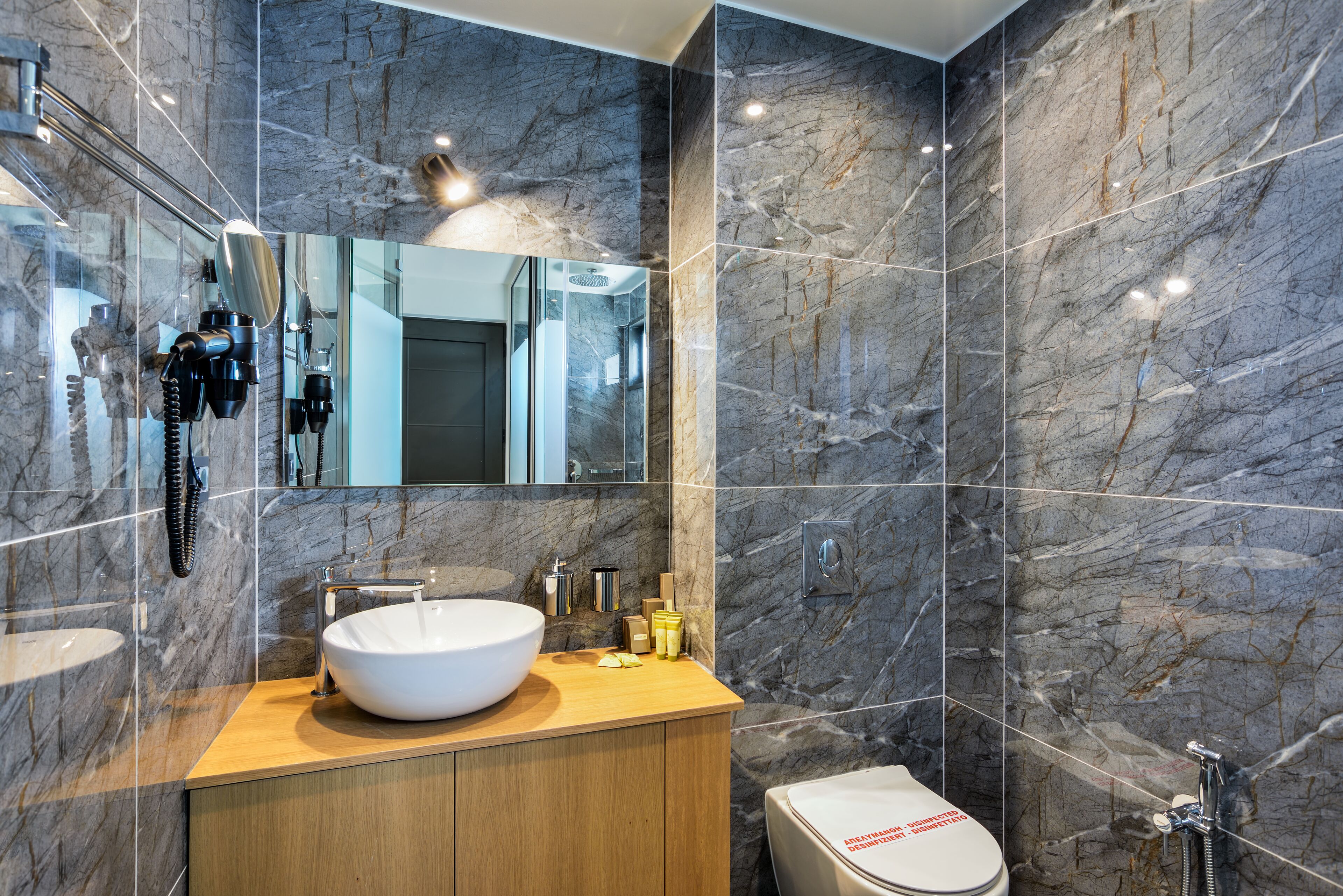 A modern bathroom with a marble wall, a wooden vanity, a white sink, and a toilet.