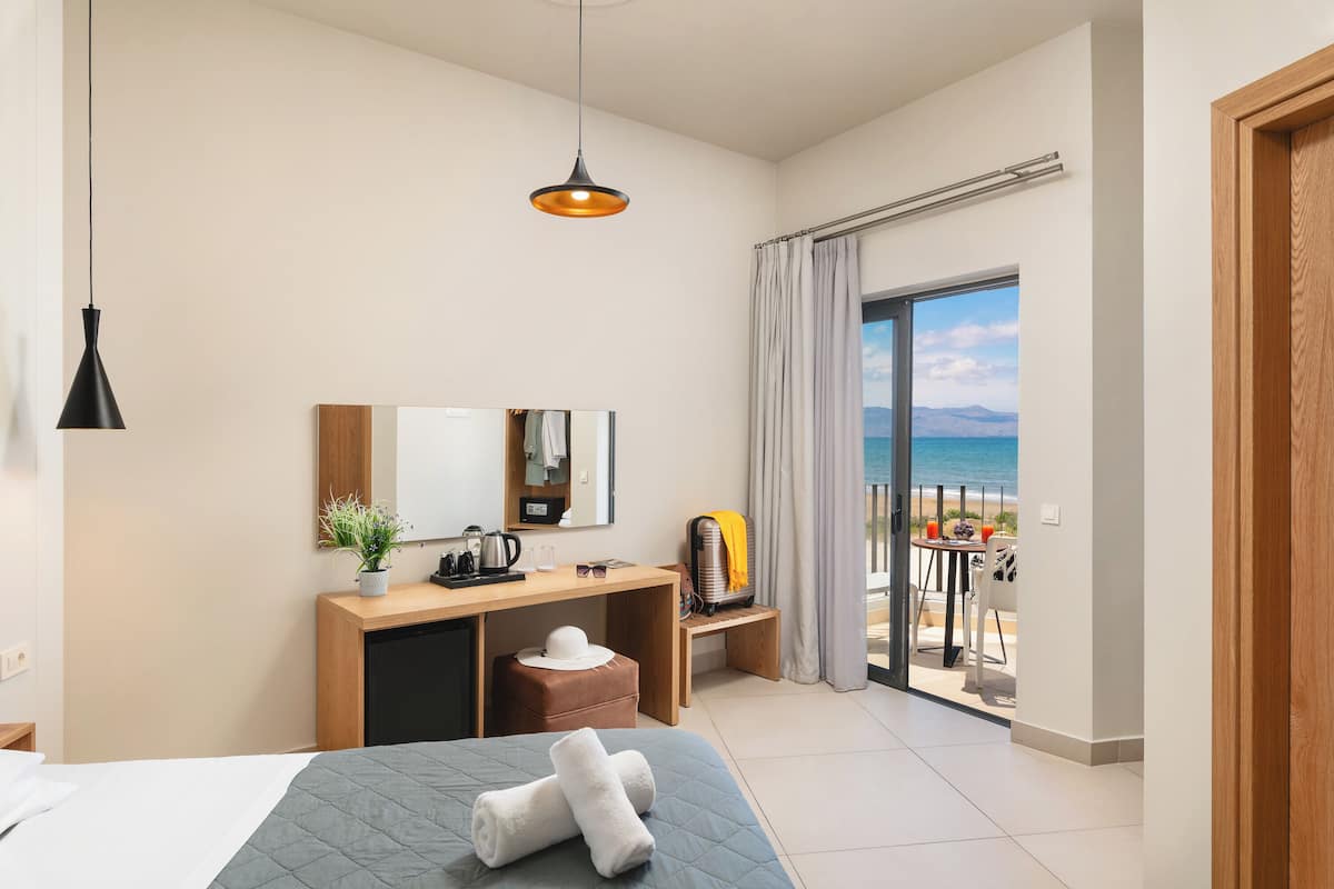 Habitación de hotel con cama, escritorio con espejo, taburete pequeño y vista a la playa a través de una puerta corrediza de vidrio.