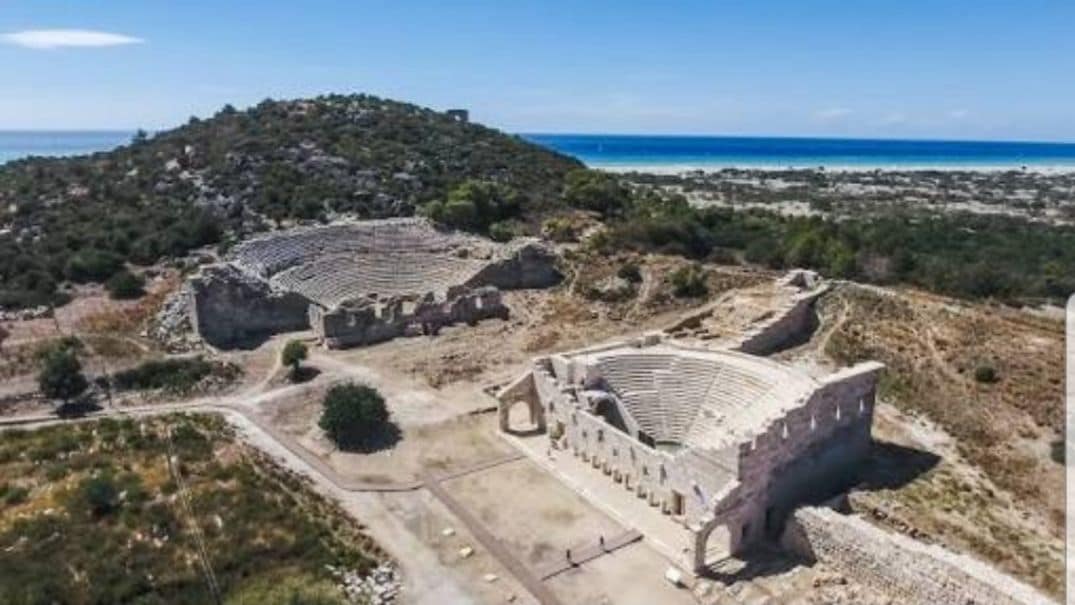 Um anfiteatro antigo com assentos em níveis e um palco central, rodeado por terreno rochoso e uma paisagem costeira.