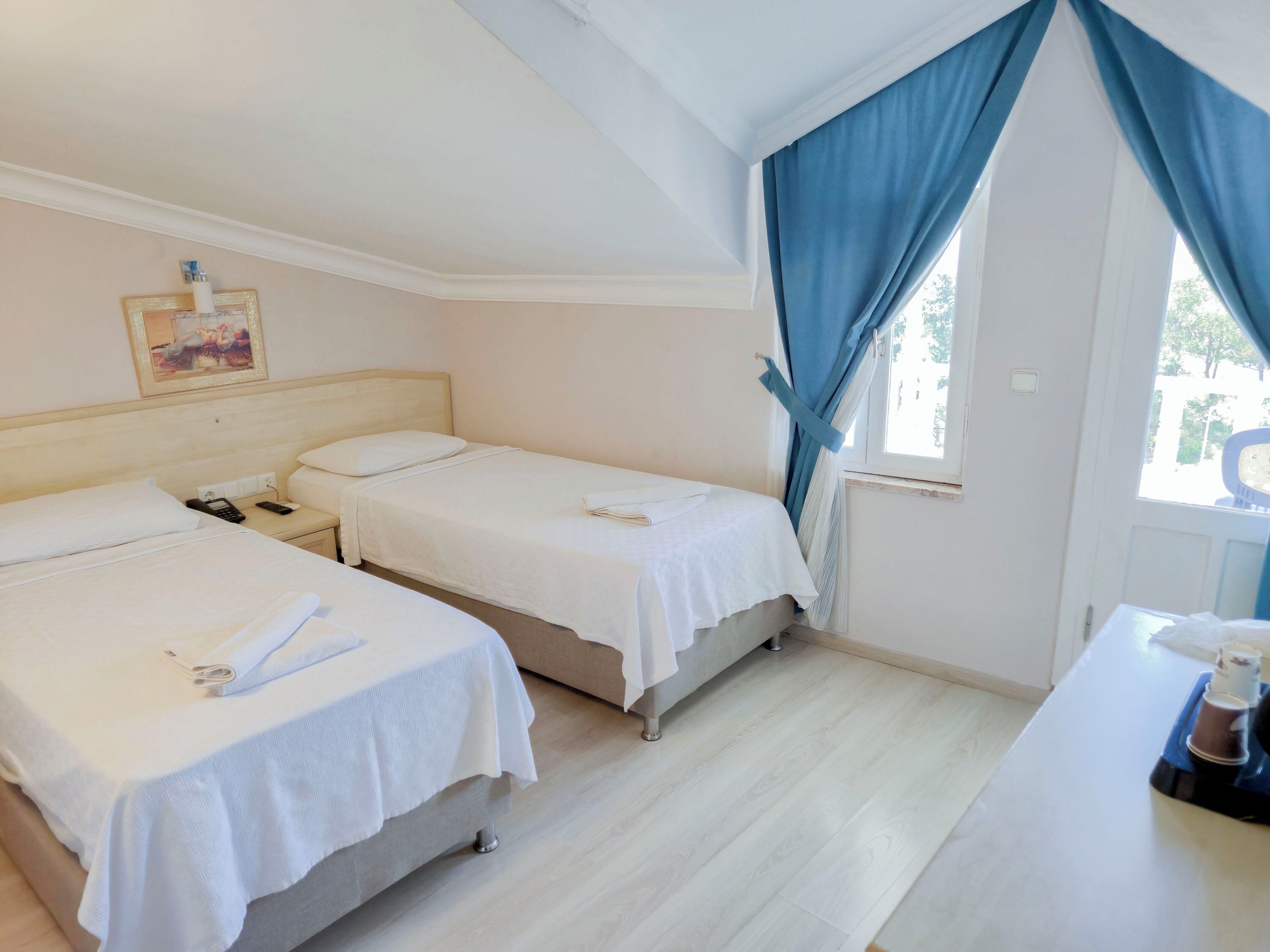 A hotel room with two single beds, a small table with a coffee maker, and a window with blue curtains.