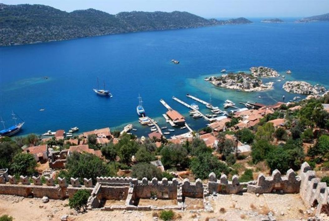 Un pueblo costero con una fortificación histórica, mar azul cristalino y barcos amarrados en el puerto.