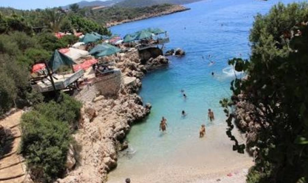 Uma área costeira com praia rochosa, água cristalina e algumas pessoas nadando. Há construções com telhados verdes, possivelmente para sombra ou abrigo.