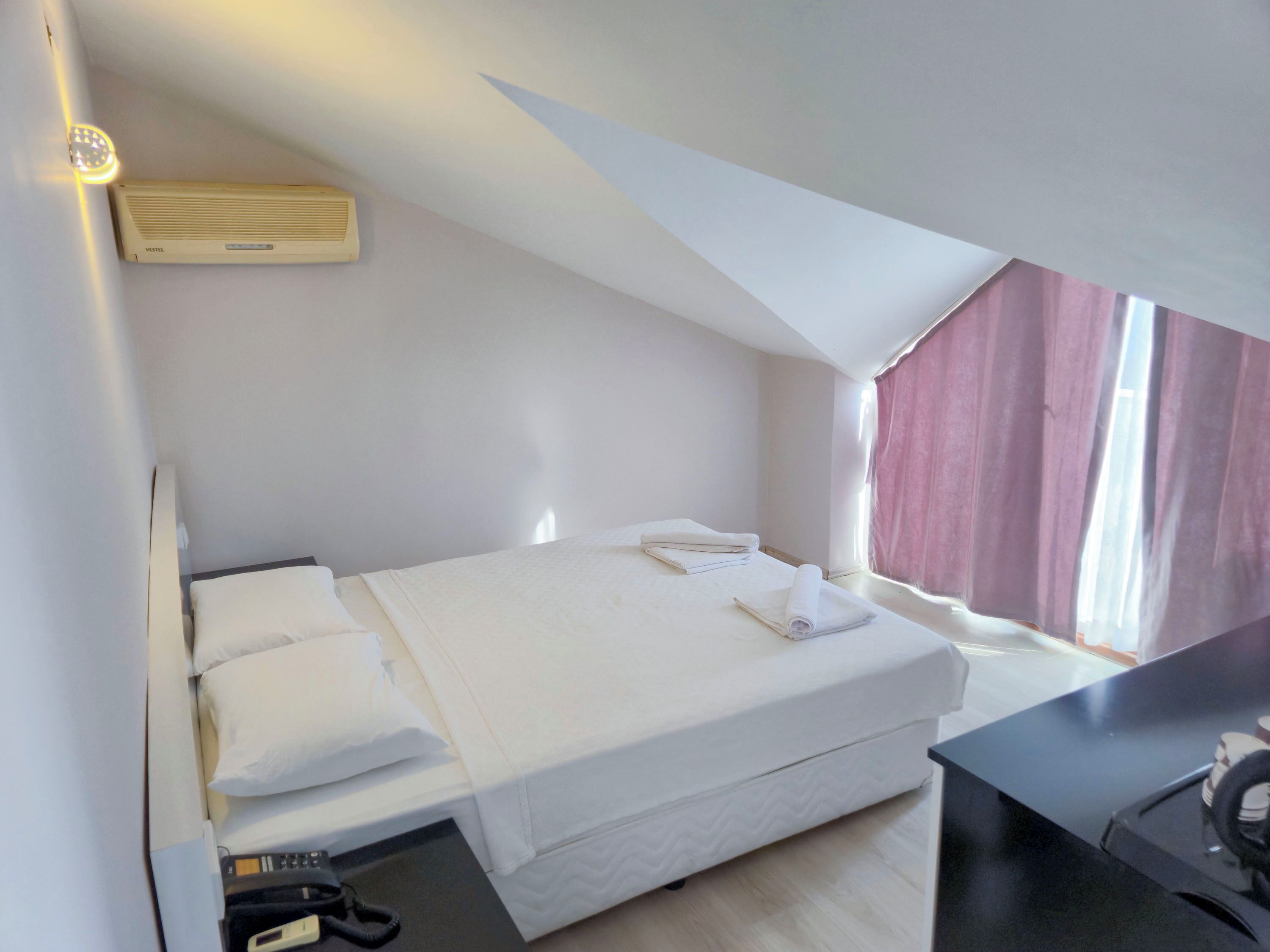 A small, well-lit room with a single bed, a desk, and a window with curtains.