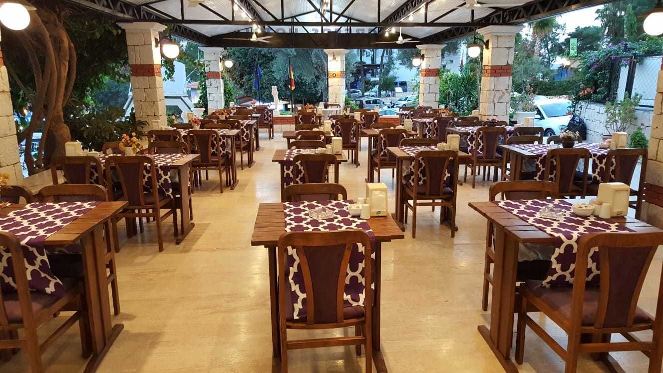 A restaurant with multiple tables and chairs, arranged in rows, with a view of the street and greenery outside.