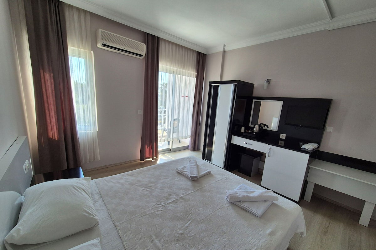 Quarto de hotel com cama, escrivaninha com espelho, televisão e janela com cortinas.