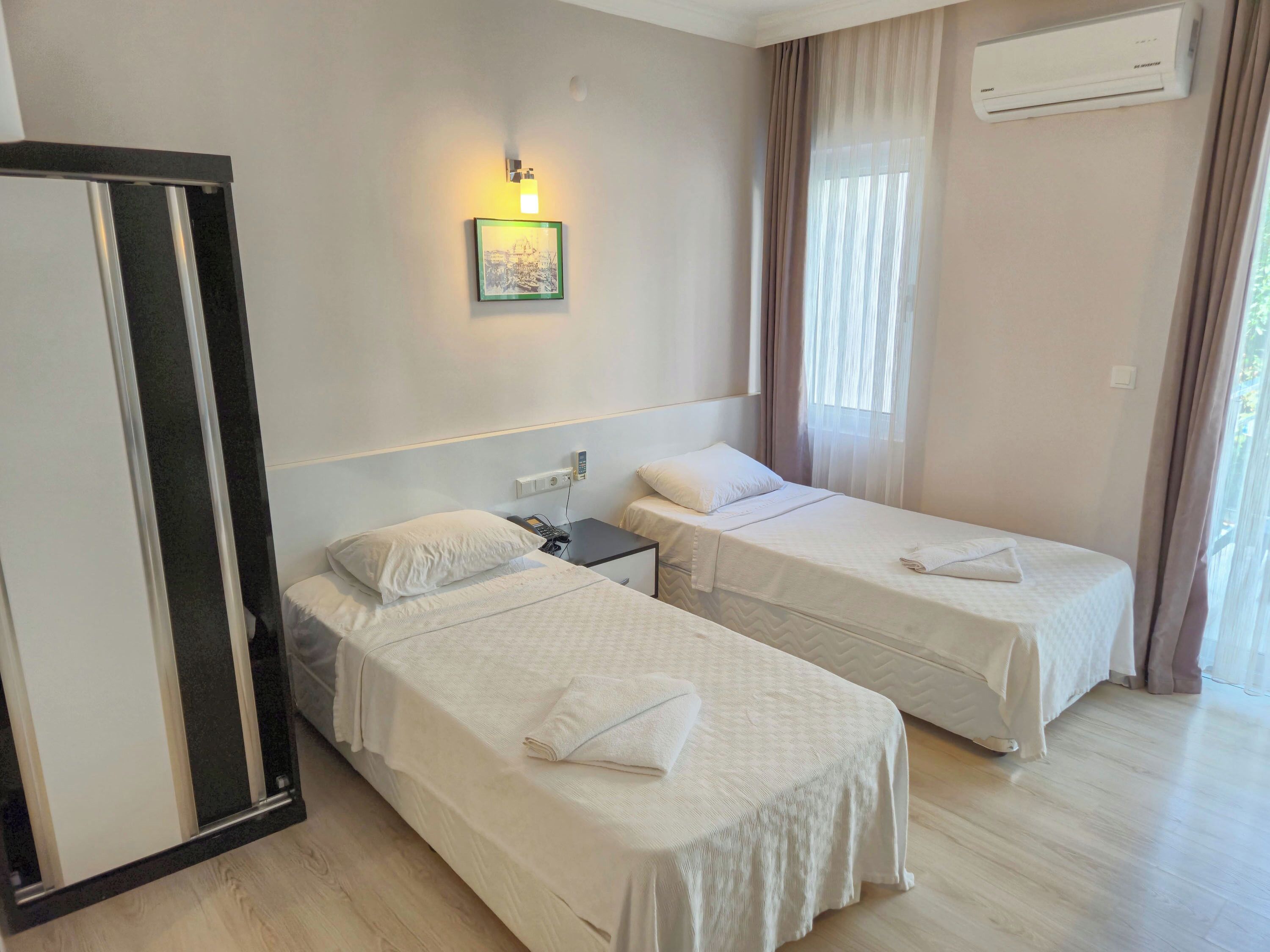 A hotel room with two single beds, a bedside table with a phone, a wall-mounted air conditioner, and a framed picture on the wall.