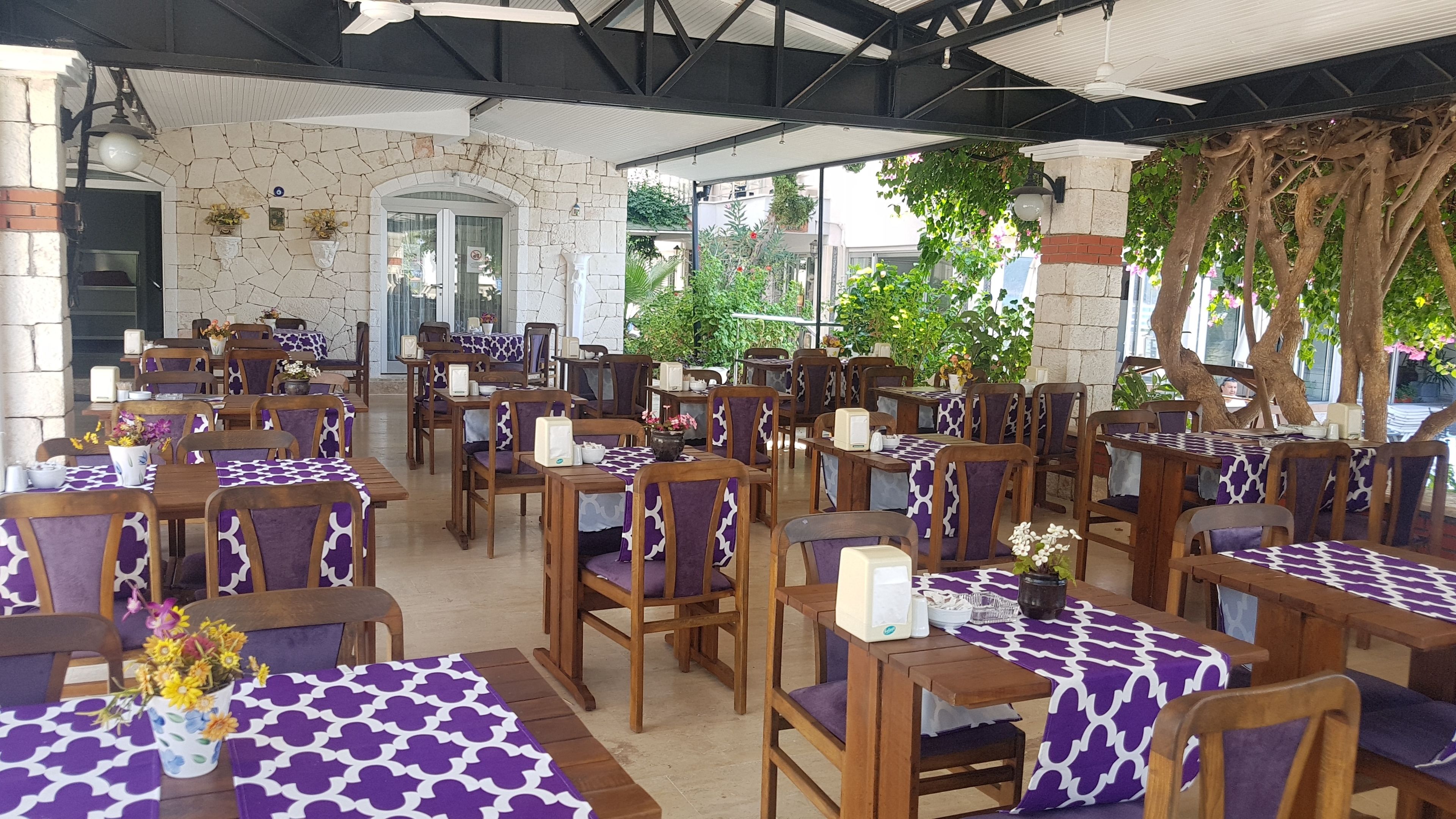 Un restaurante con terraza, paredes de piedra y abundante vegetación. Las mesas están cubiertas con manteles morados y blancos, y las sillas tienen cojines morados.