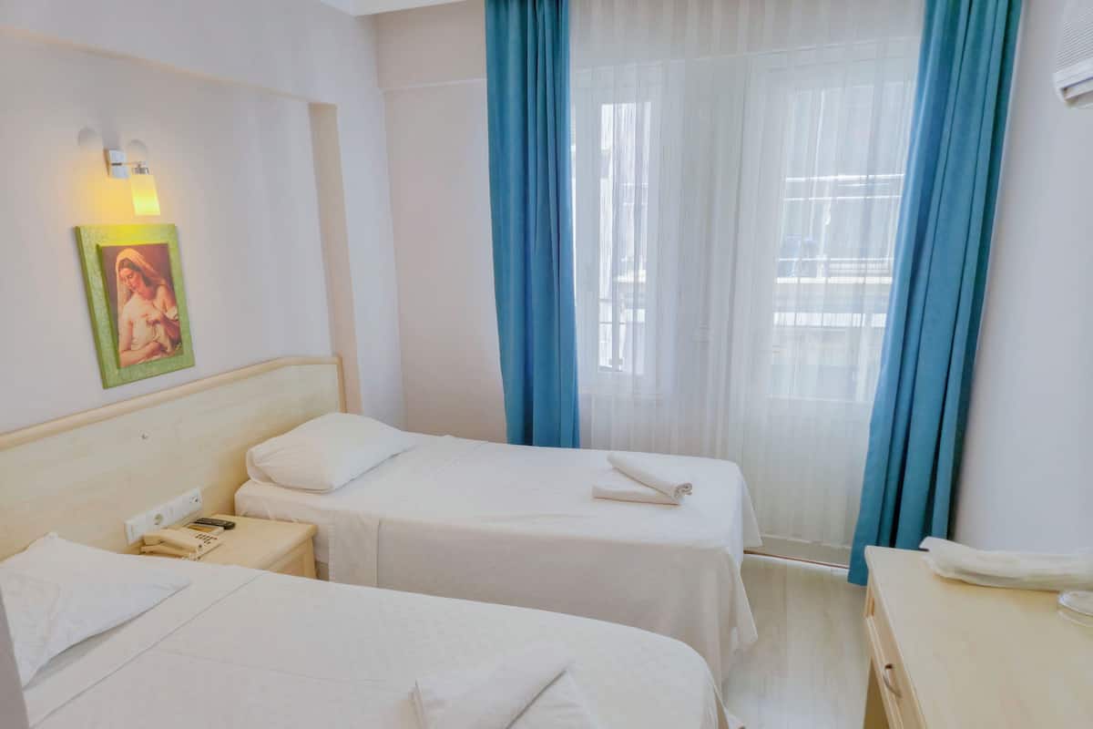 Quarto de hotel com duas camas, uma mesa de cabeceira com telefone, uma luminária na parede e uma janela com cortinas azuis.