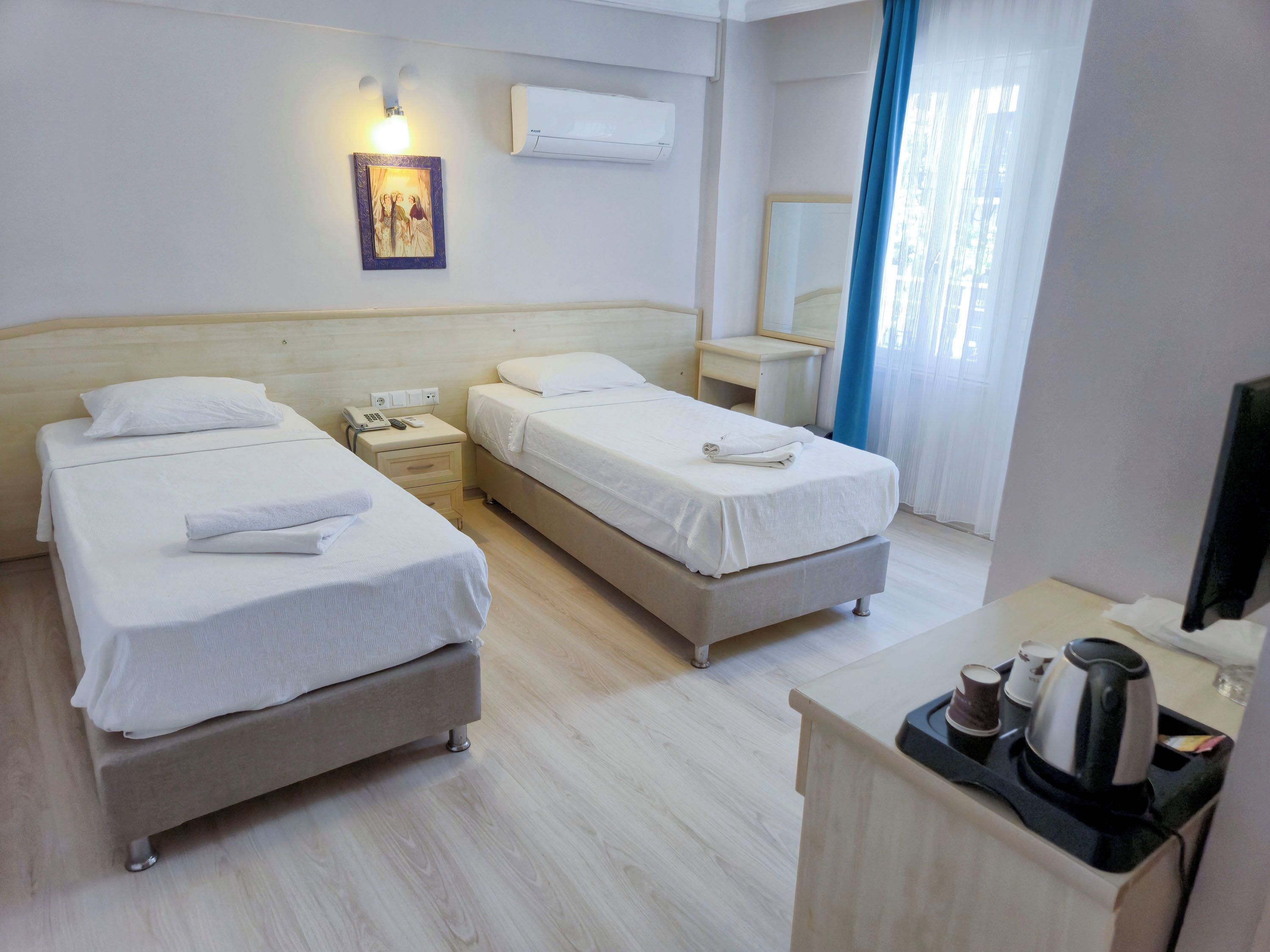 A hotel room with two beds, a bedside table with a phone and a lamp, a desk with a mirror, and a window with curtains.