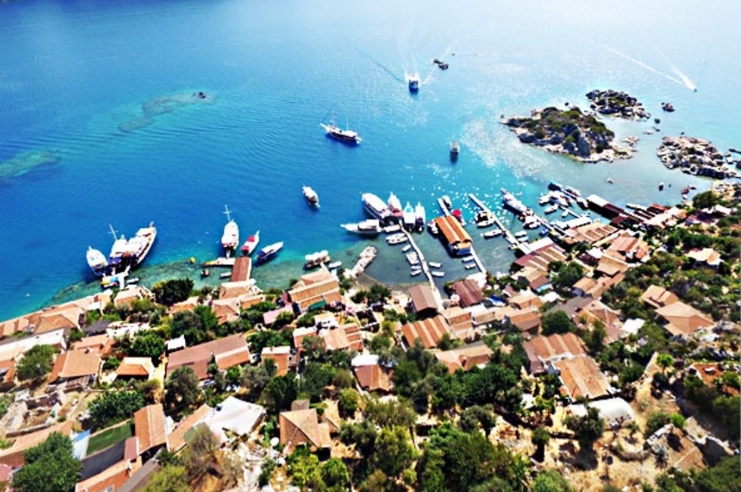 An aerial view of a coastal town with boats docked at a marina, surrounded by residential buildings and greenery.