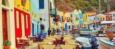 Una calle costera con edificios coloridos, mesas al aire libre y barcos amarrados al agua.