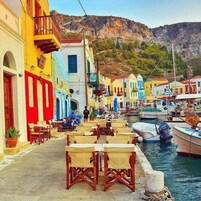 A coastal street with colorful buildings, outdoor seating, and boats docked along the water.