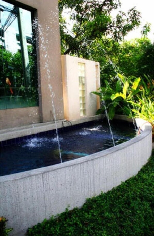 A rectangular water feature with two streams of water, surrounded by greenery and a building with a glass facade. - Tongtip Place (Bangkok)