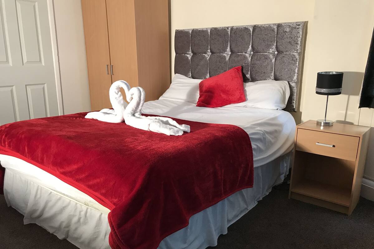 A neatly made bed with a red blanket, a white pillow, and a red pillow. There is a wooden nightstand with a lamp beside the bed.