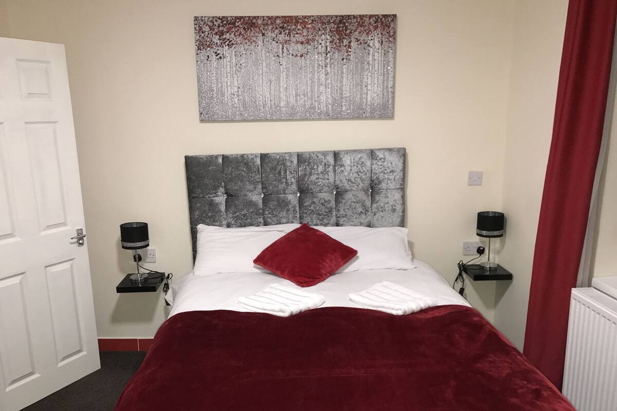 A neatly made bed with a red throw pillow, a grey tufted headboard, and two bedside tables with lamps.