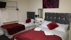 A hotel room with two beds, each with a red blanket and white linens. There is a television on the wall, a mirror, and a painting of a rose. - Hello Roomz (Blackpool)