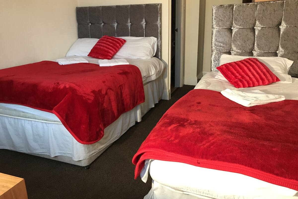 Two single beds with grey headboards, red blankets, and white pillows in a room with a carpeted floor.