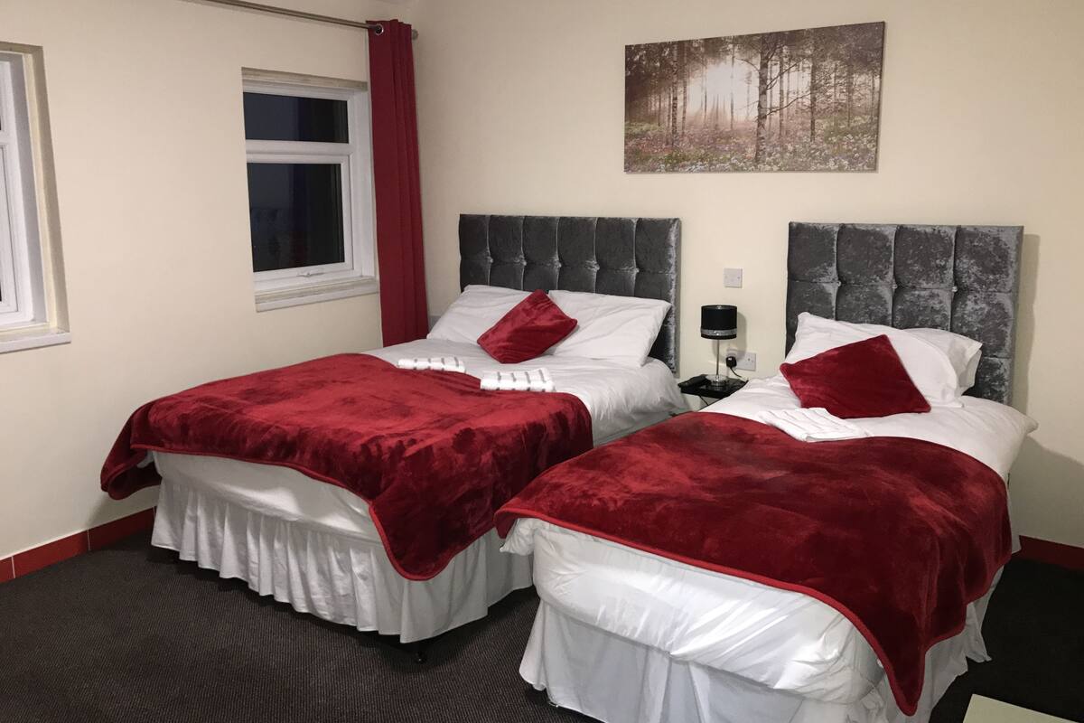 A hotel room with two beds, each with a red bedspread and white pillows, a grey headboard, a window with a red curtain, and a painting of a forest on the wall.