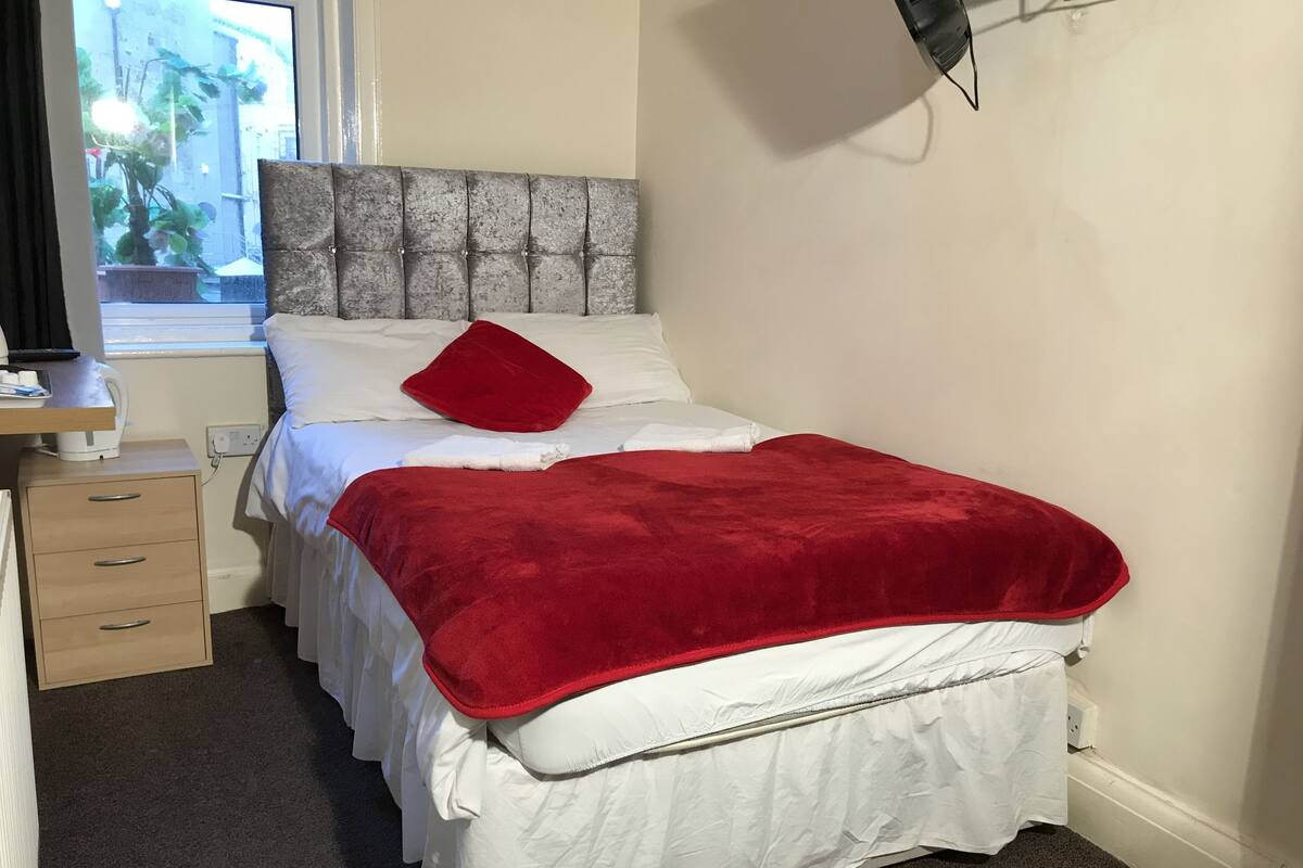 A neatly made bed with a red blanket, white bedding, and a grey headboard. There is a wooden nightstand with a lamp and a power outlet.