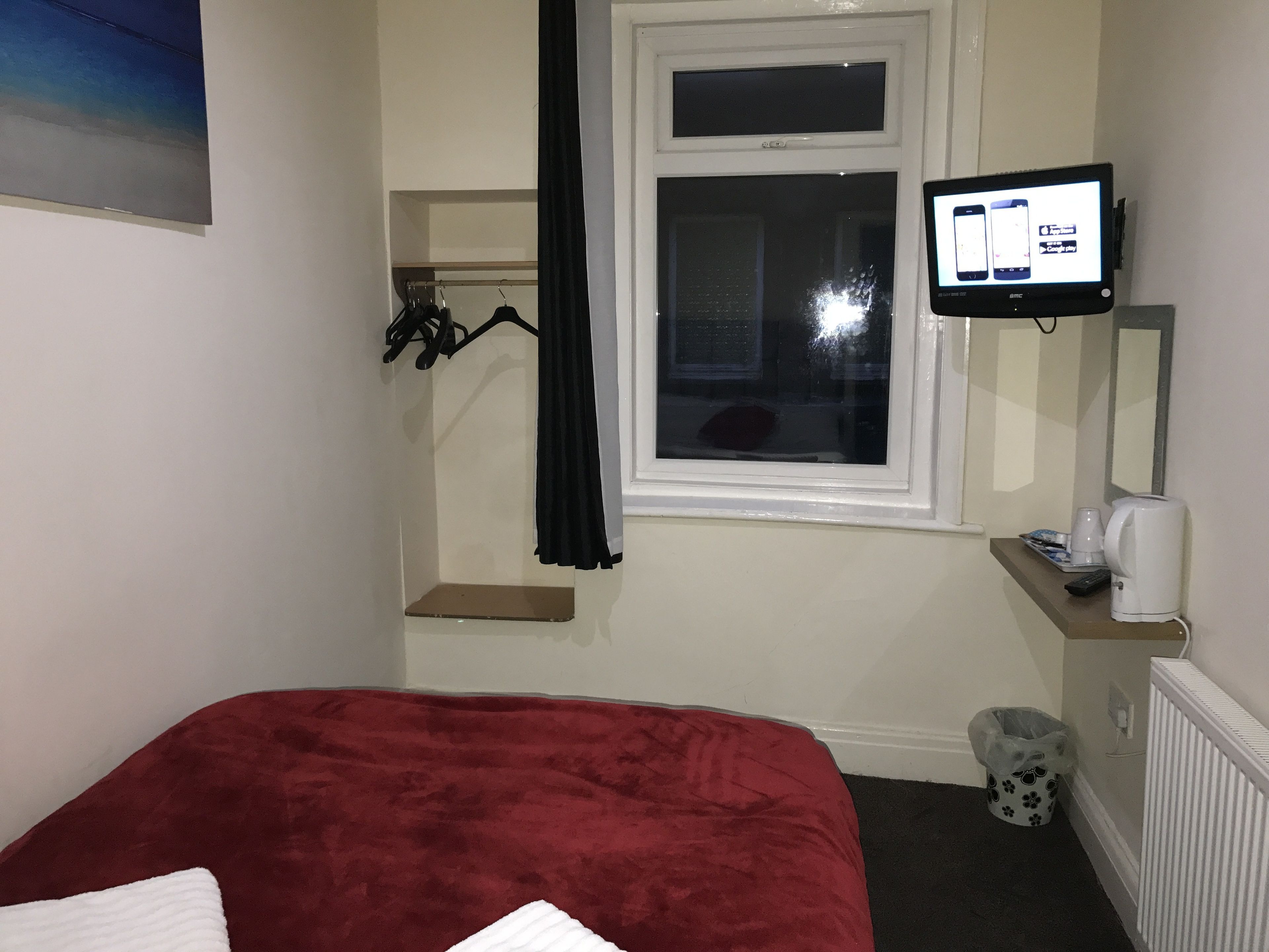 A hotel room with a bed, a TV mounted on the wall, a window with a curtain, a small shelf with a coffee maker, and a trash bin.