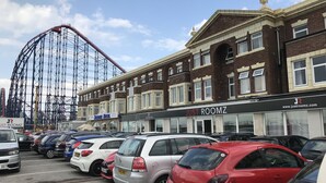 A parking lot with cars, a building with a sign for 'JUST ROOMZ', and a roller coaster in the background. - Hello Roomz (Blackpool)