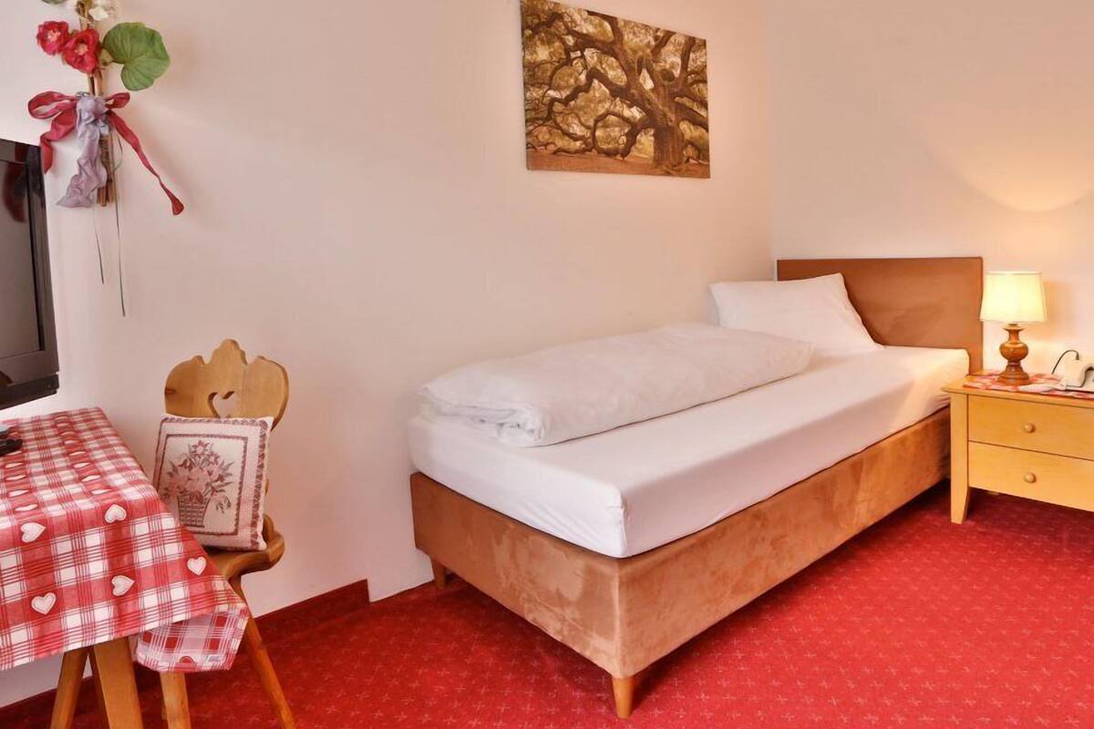 A neatly arranged hotel room with a single bed, a bedside table with a lamp, a small table with a checkered tablecloth, and a wall-mounted artwork.