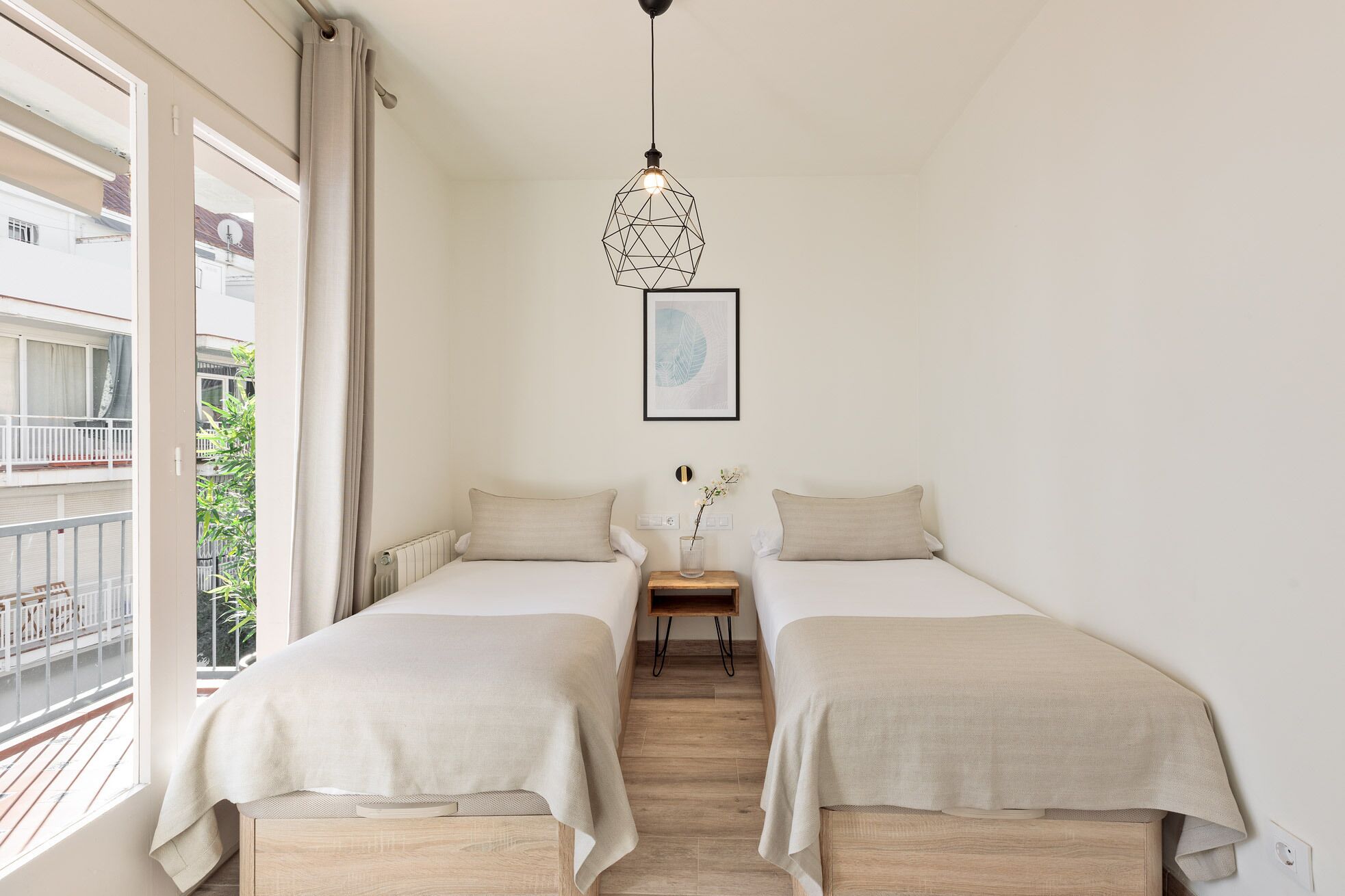 A bedroom with two beds, a wooden floor, a window with a view of a balcony, and a hanging light fixture.