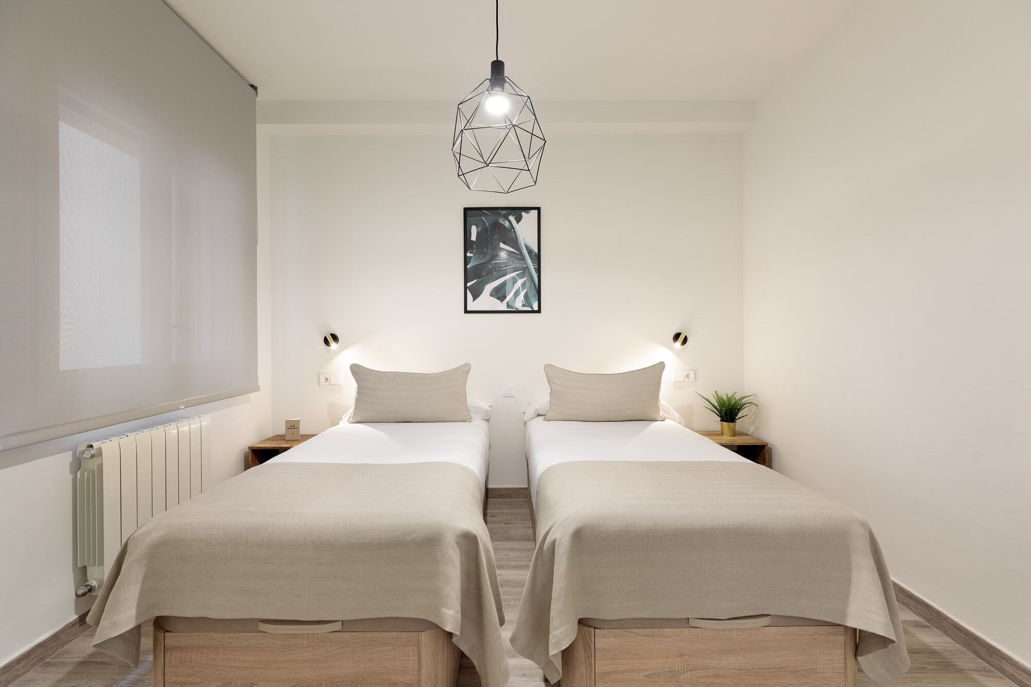 A hotel room with two beds, a geometric pendant light, a framed abstract artwork, bedside tables with lamps, and a radiator.