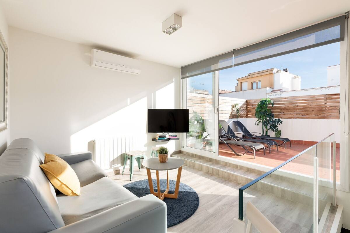 A modern living room with a sofa, a TV, a small table, and a balcony with a chair and plants.