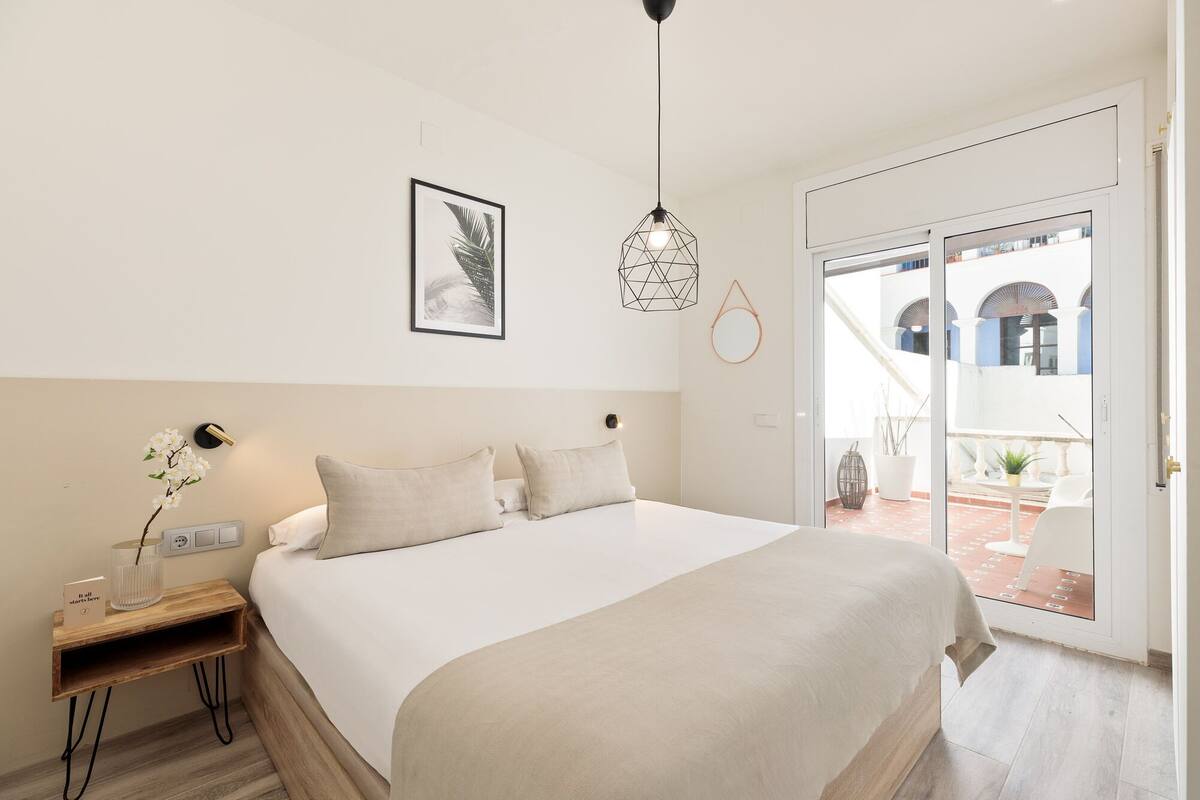 A bedroom with a bed, a nightstand, a wall-mounted lamp, a framed picture, and a view of the outside balcony.