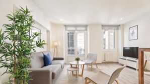 Un salon moderne avec un canapé, des fauteuils, une table basse et un téléviseur. On y trouve également une grande plante et un lampadaire.