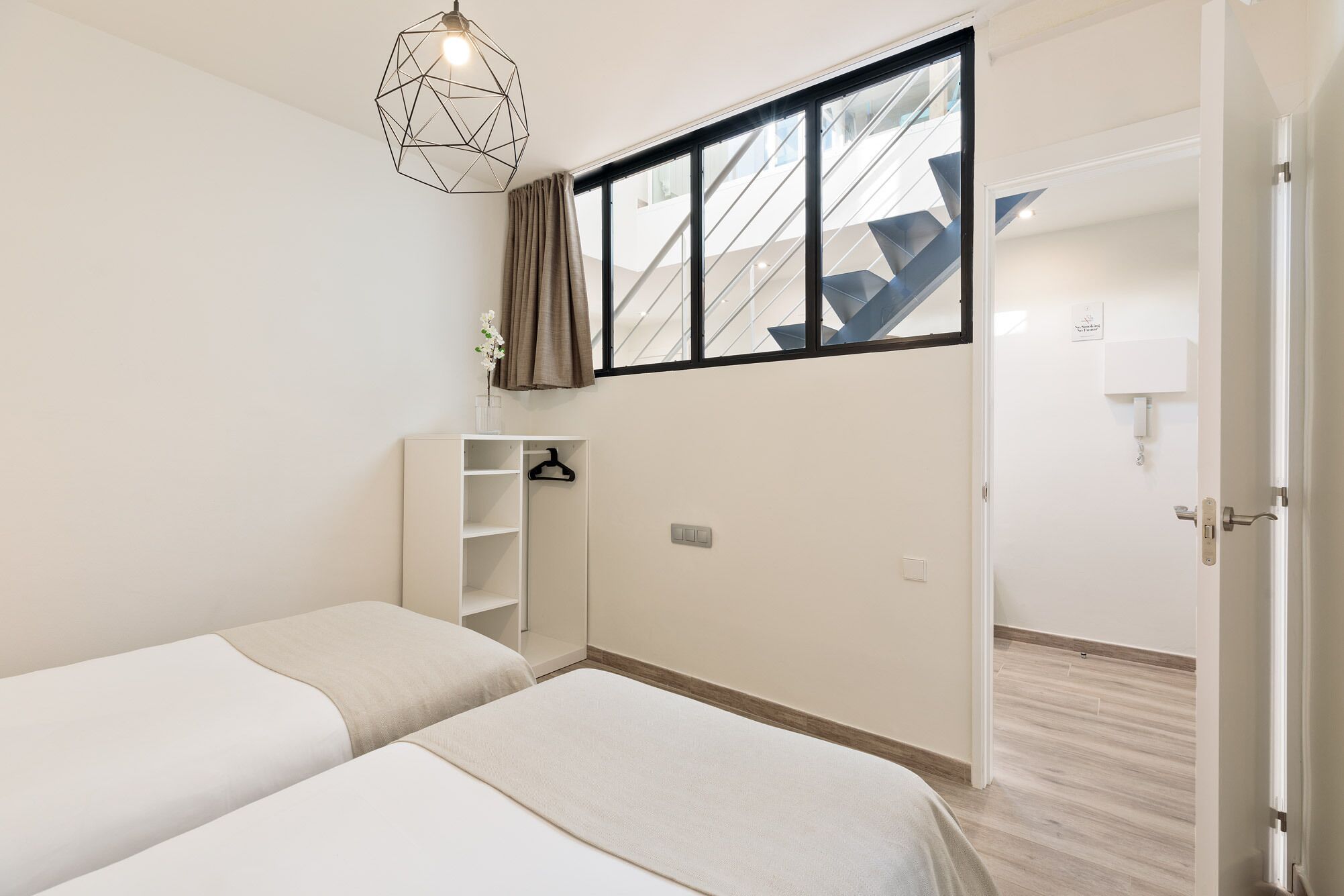 A minimalist bedroom with two beds, a wardrobe, and a window with a view of a staircase.