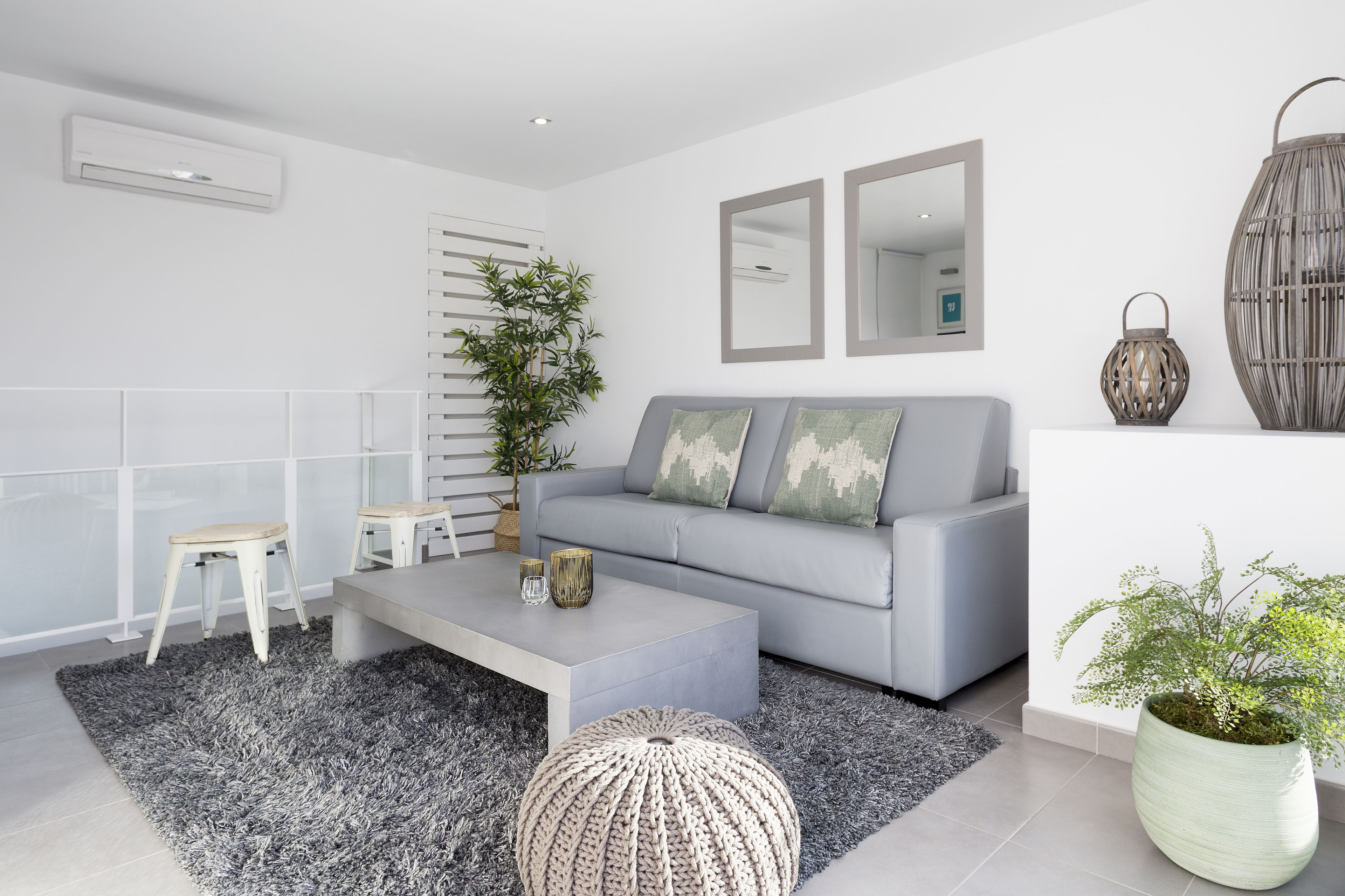 Un salon moderne avec un canapé gris, deux tabourets blancs, une table basse grise et une grande plante en pot.