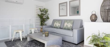 Un salon moderne avec un canapé gris, deux tabourets blancs, une table basse grise et une grande plante en pot.